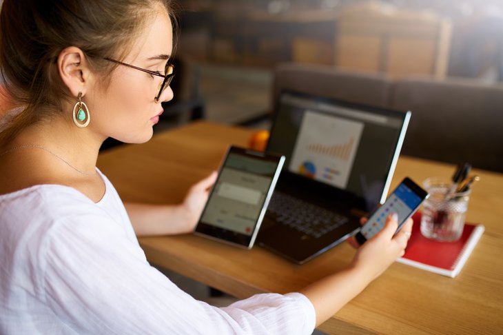 Une jeune femme à lunettes travaille à une table avec un smartphone, une tablette et un ordinateur portable