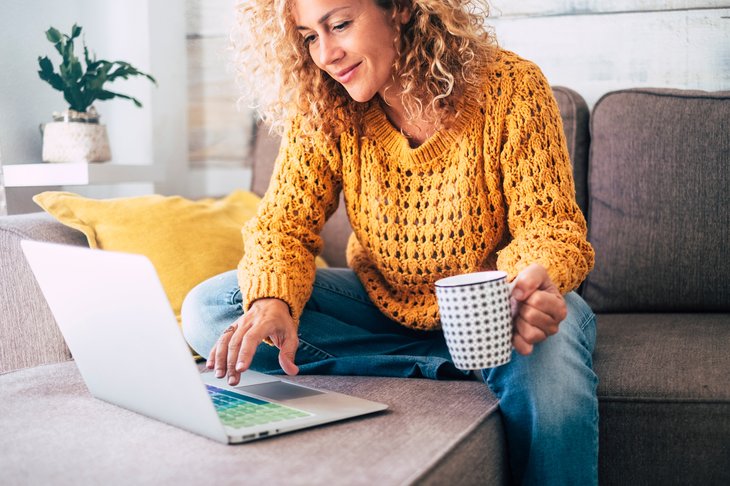 Une femme boit dans une tasse de café et tape sur le clavier d'un ordinateur portable alors qu'elle est assise sur le canapé de son salon à la maison