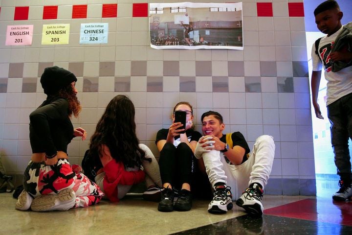 Students sit and kneel on the floor of a school hallway while looking at their phones.